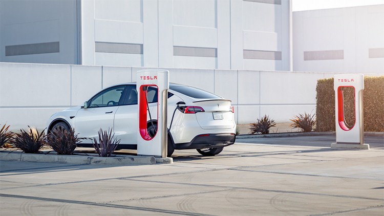 Weißes Elektroauto lädt an einer Ladestation mit rotem Tesla-Logo auf einem Parkplatz neben niedrigem Pflanzenbewuchs.