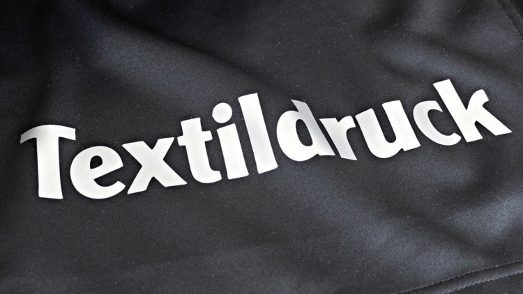Das Wort „Textildruck“ ist in weißer Schrift auf einem schwarzen Stoffhintergrund abgebildet.
