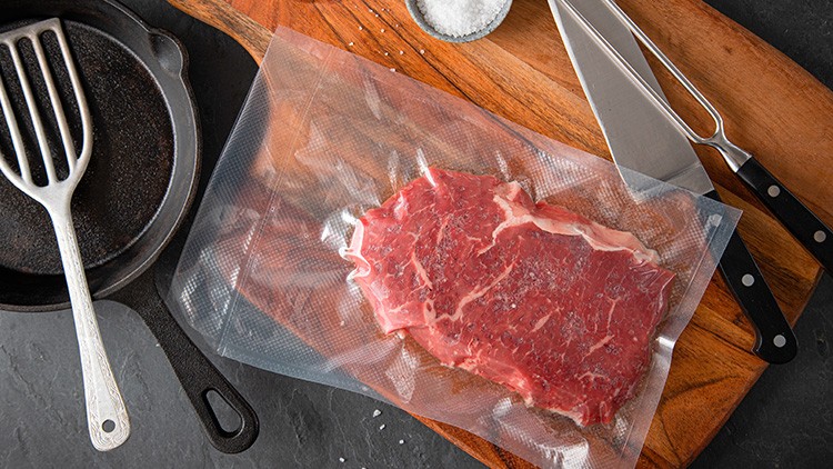 Rotes vakuumverpacktes Steak liegt auf einem Holzbrett, daneben Pfanne, Salzschale, Messerset und Grillwender.