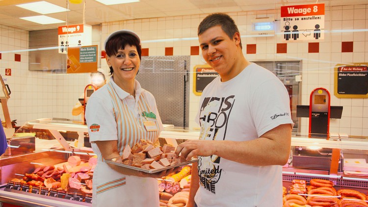 Mitarbeiterin in weißer Arbeitskleidung reicht Tablett mit Fleischproben vor einer Wurst-Theke an eine Person weiter.