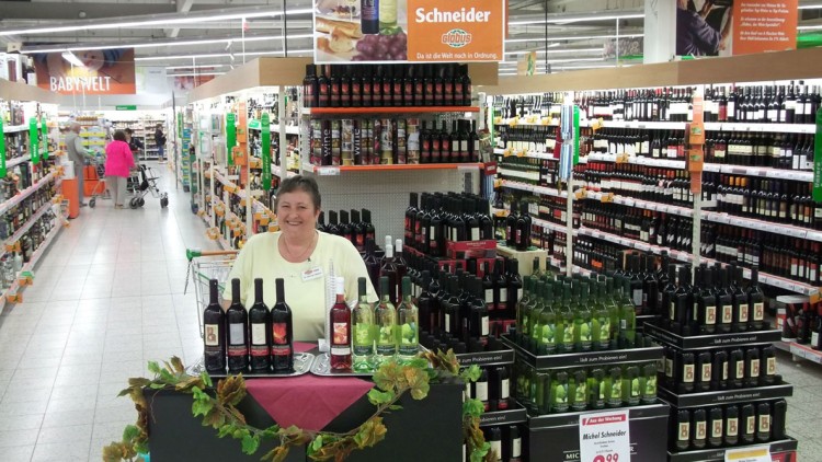 Verkostungsstand mit Weinflaschen vor einem Regal in der Weinabteilung, darüber ein Schild mit „Weine von Michael Schneider“.