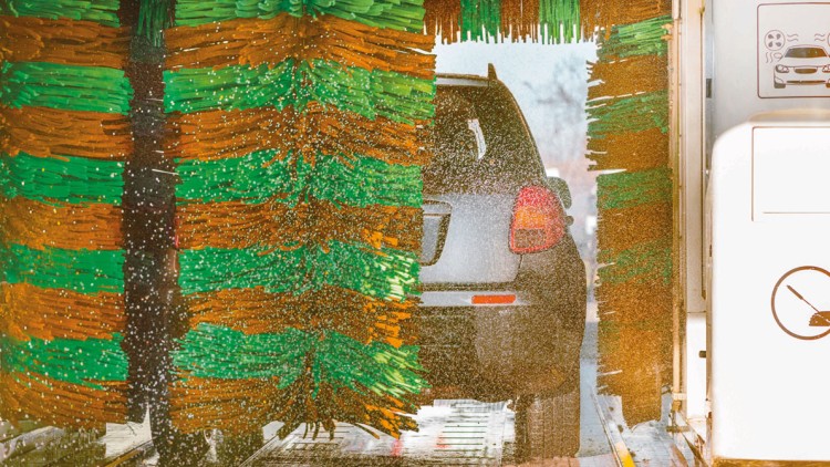 Ein graues Auto wird in einer Waschanlage von rotierenden Bürsten in Grün und Orange mit Wasser besprüht.