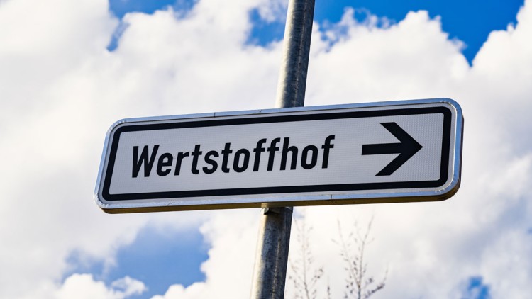 Ein Schild mit der Aufschrift Wertstoffhof und einem Pfeil nach rechts steht vor einem Himmel mit weißen Wolken.