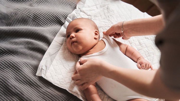 Ein Baby liegt auf einer weißen Unterlage, während zwei Hände das weiße Oberteil des Babys richten.