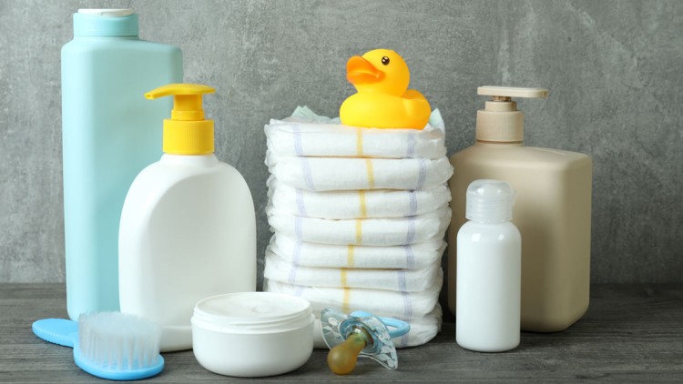 Mehrere Babyartikel wie Lotionen, Bürste, Creme, Schnuller, Windeln und eine gelbe Gummiente vor grauem Hintergrund.