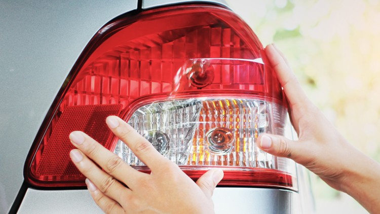 Zwei Hände berühren ein rotes und weißes Rücklicht an einem silbernen Auto vor verschwommenem Hintergrund mit Tageslicht.