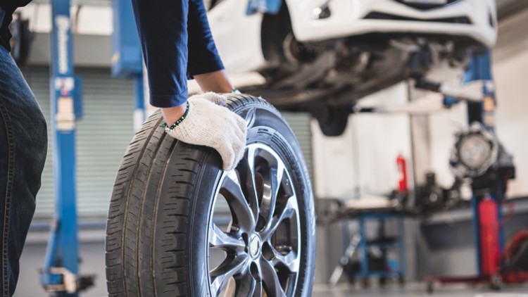 Person mit hellen Handschuhen hält ein Autoreifen neben einer Hebebühne mit einem angehobenen weißen Auto in einer Werkstatt.
