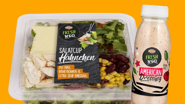Eine Verpackung mit Salatcup Hähnchen und eine Flasche Fresh N'Go American Dressing vor einem orangefarbenen Hintergrund.