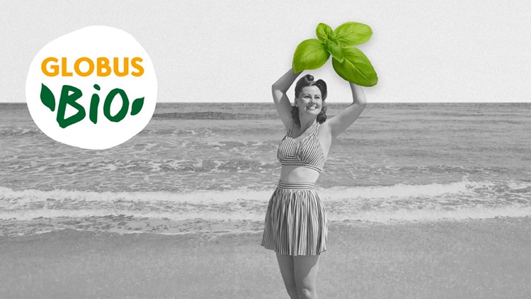 Person in gestreifter Kleidung steht am Strand, hält große Basilikumblätter über dem Kopf, Logo GLOBUS Bio links oben.