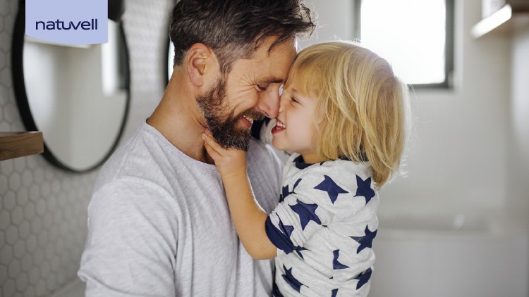 Ein Kind mit blondem Haar und Sternenmuster-Pullover wird von einer Person im grauen Oberteil auf dem Arm gehalten, Logo „natuvell“.