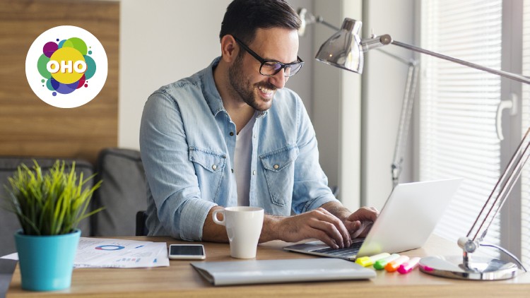 Person in Jeanshemd arbeitet an einem Laptop an einem Schreibtisch mit Kaffeetasse, Pflanzen, Stiften und Unterlagen, OHO-Logo links.