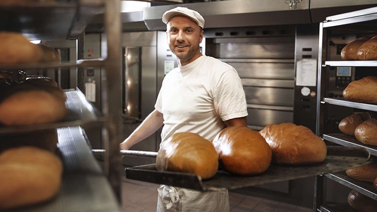 Person im weißen T-Shirt und weißer Mütze hält ein Blech mit frisch gebackenen Broten in einer Bäckerei vor Öfen.