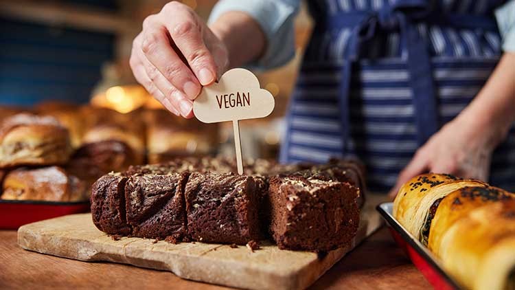 Eine Hand platziert ein Schild mit der Aufschrift „VEGAN“ in Stücke eines Schokokuchens auf einem Holzbrett.