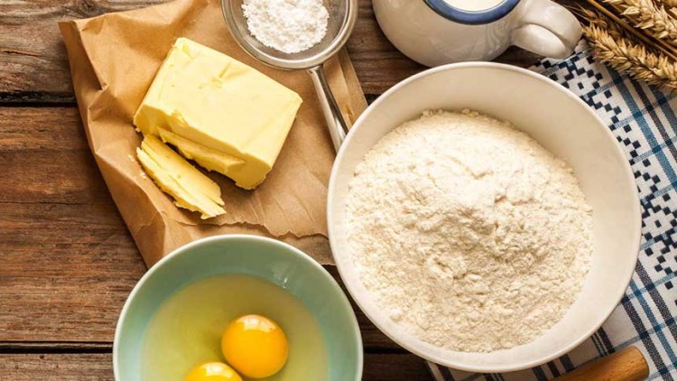 Ein Holzuntergrund mit Butter, Mehl, zwei Eiern in Schale, Milchkanne, Zucker und Weizenähren liegt bereit.