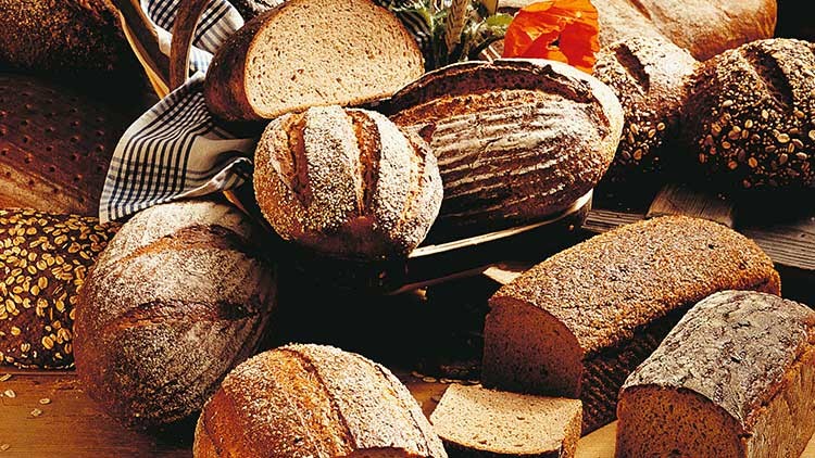 Verschiedene Sorten frisch gebackenes Brot, darunter Vollkorn- und Roggenbrote, teils angeschnitten, auf einem Holztisch.