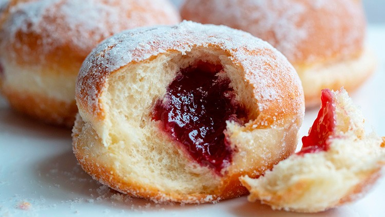 Ein mit Puderzucker bestreuter Berliner mit roter Marmeladenfüllung, daneben ein kleines abgebissenes Stück Teig.