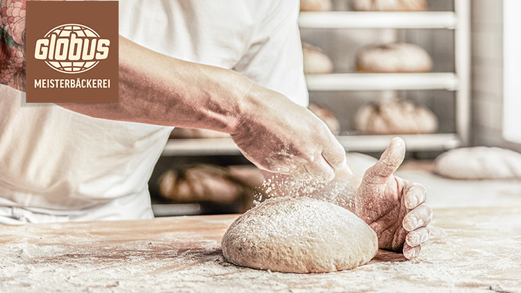 Ein Bäcker formt einen mit Mehl bestäubten Brotteig auf einem Holztisch, im Hintergrund sind Brotlaibe und Regale zu sehen.