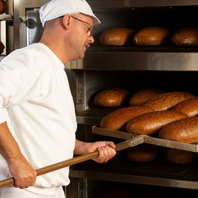 Eine Person in weißer Arbeitskleidung holt mit einem Backschieber frisch gebackene Brote aus einem großen Backofen.