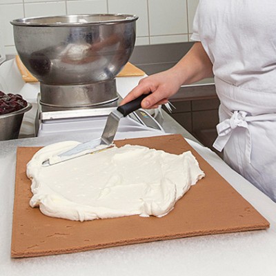 Eine Person in weißer Arbeitskleidung verteilt mit einer Winkelpalette Creme auf einem braunen Kuchenboden, links stehen Schüsseln.