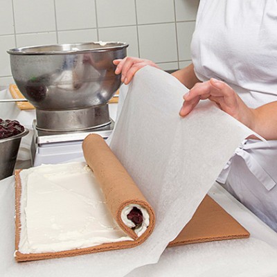 Eine Person in weißer Kleidung rollt eine gefüllte Teigrolle mit Sahne und Kirschen auf Backpapier in einer Küche ein.