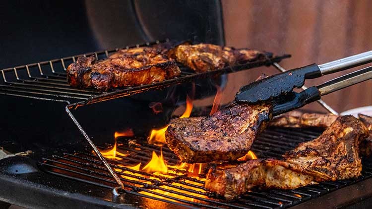 Mehrere Fleischstücke liegen auf einem Grill mit offenen Flammen, während eine Grillzange ein Steak anhebt.