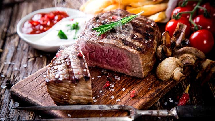 Ein Medium-rare gegrilltes Steak mit Salz und Kräutern liegt neben Pilzen, Tomaten, Pommes und Dips auf einem Holzbrett.
