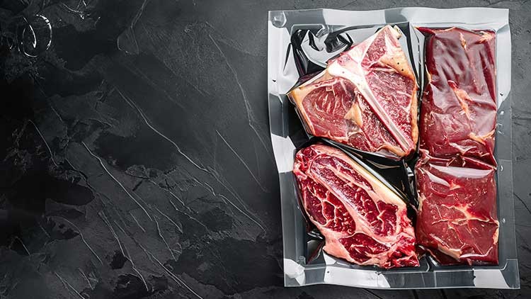 Vier vakuumverpackte Fleischstücke liegen nebeneinander auf einer dunklen, leicht strukturierten Steinoberfläche.