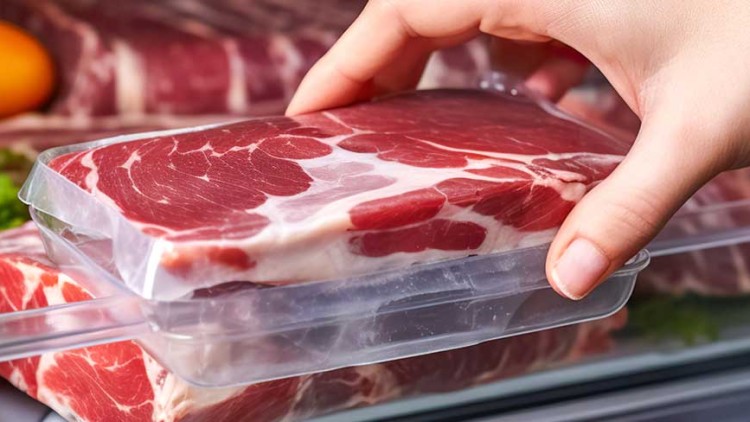 Hand nimmt eine transparente Verpackung mit rohem Fleisch aus einem Kühlschrank, dahinter liegen Gemüse und weitere Fleischstücke.