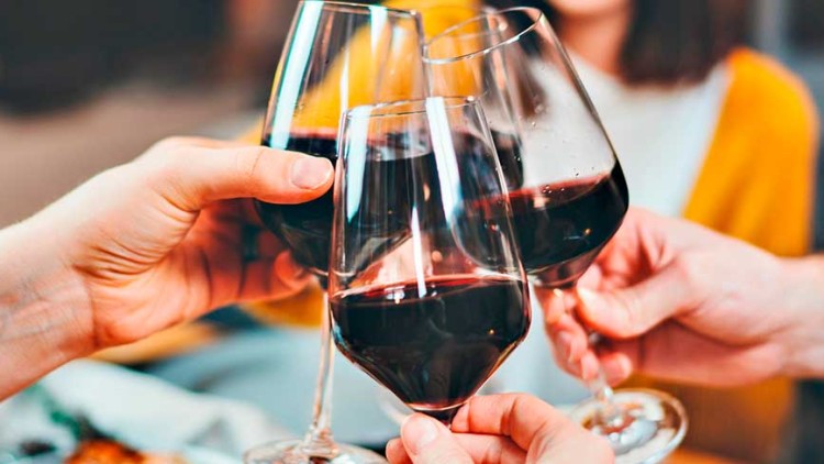 Vier gefüllte Weingläser mit Rotwein stoßen an, Hände halten die Gläser, unscharfer Hintergrund zeigt Personen und Tischdekoration.