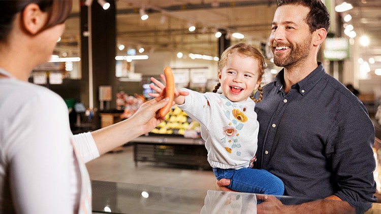 Eine erwachsene Person überreicht einem Kind eine Wurst, das von einer weiteren Person auf dem Arm gehalten wird, in einem Supermarkt.