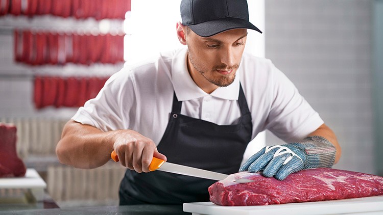 Eine Person im weißen Polo und schwarzer Schürze schneidet mit einem Messer ein großes Stück rohes Fleisch auf einem Schneidbrett.