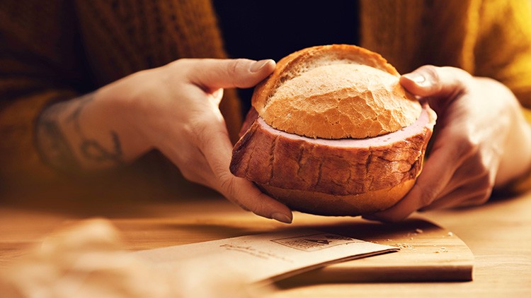 Eine Person hält ein aufgeschnittenes Brötchen mit einer dicken Scheibe Fleischwurst über einem Tisch mit einem Papierbrief.