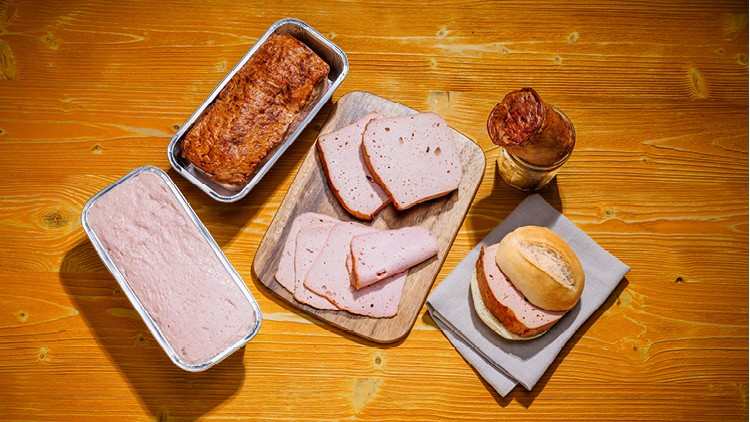 Verschiedene Varianten von Fleischkäse, geschnitten und als ganzer Laib, auf Holzuntergrund mit Brötchen und Aufschnitt.