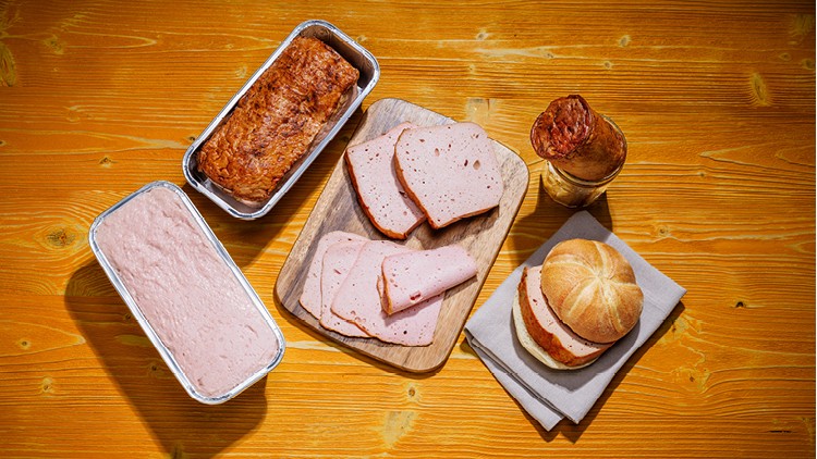 Verschiedene Varianten von Fleischkäse auf Holzuntergrund, darunter Scheiben, Brötchen, gebackene und ungebackene Zubereitungen.