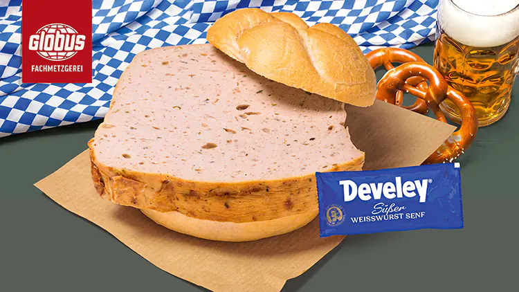 Ein Leberkäsbrötchen mit Senfbeutel der Marke Develey, daneben eine Brezel, ein Bierkrug und ein blau-weißes Tuch.