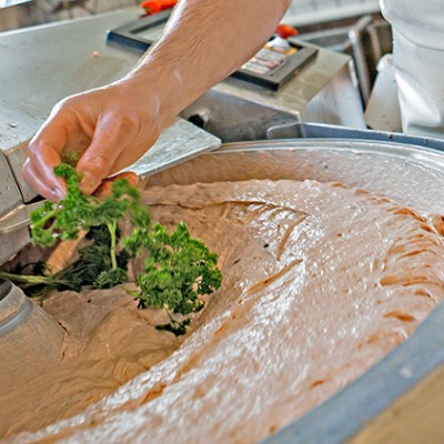 Eine Hand gibt frische Petersilie in eine große industrielle Schüssel mit Fleischteig in einer Küchenmaschine.