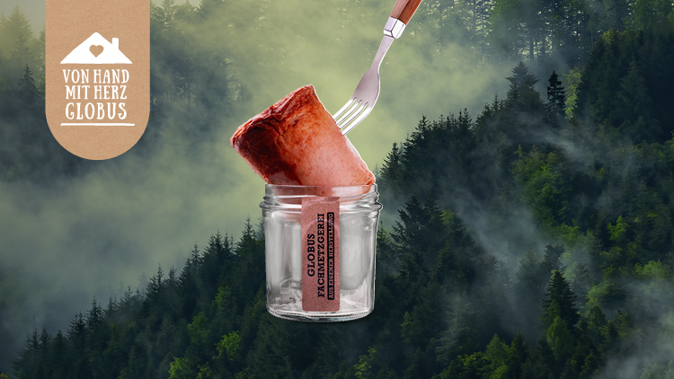 Ein Glas mit der Aufschrift „GLOBUS Fachmetzgerei“ und eine Gabel mit einem Fleischstück schweben vor einem nebligen Waldhintergrund.