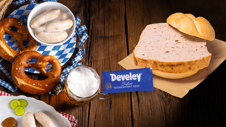 Auf einem Holztisch liegen Weißwürste, Brezeln, Bier, Leberkäse mit Brötchen und Develey Süßer Weißwurst Senf.