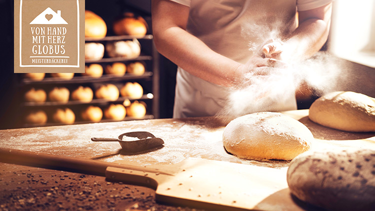 Eine Person mehlt Hände über einem Arbeitstisch mit Brotteig, im Hintergrund ein Regal mit goldbraunen Broten, Symbol mit Text: Von Hand mit Herz GLOBUS Meisterbäckerei.