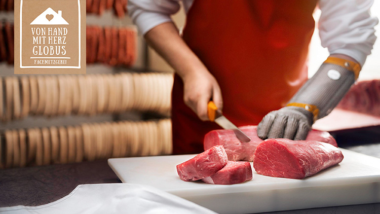 Ein Mann mit Kettenhandschuh schneidet rotes Rindfleisch auf einem weißen Schneidebrett, links ist ein Logo mit „Von Hand mit Herz GLOBUS Fachmetzgerei“.