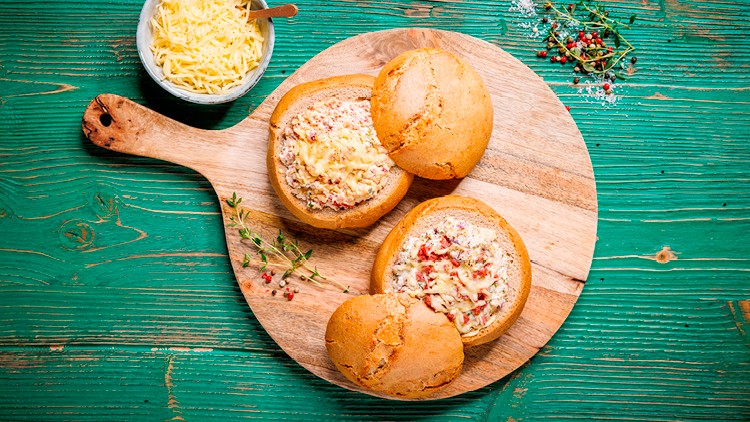 Drei gefüllte Brotlaibe mit Käse überbacken liegen auf einem Holzbrett, daneben Kräuter und eine Schale mit geriebenem Käse.