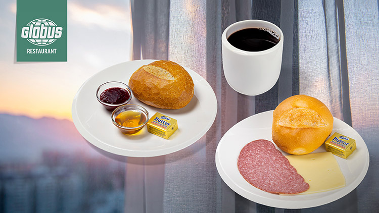 Zwei Frühstücksteller mit Brötchen, Butter, Salami, Käse, Marmelade, Honig und Kaffee vor einem Fenster mit Vorhängen im Hintergrund.