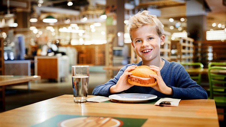 Kind in blauem Pullover sitzt an einem Holztisch und hält einen Burger, daneben ein Glas Wasser und ein Teller.