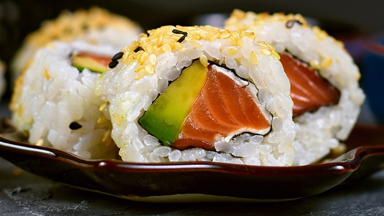 Nahaufnahme von Sushi-Rollen mit Lachs, Avocado, Reis, Sesam und schwarzem Sesam auf einem dunklen Teller.