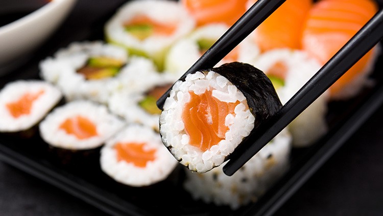 Ein Sushi-Rollstück mit Lachsfüllung wird von schwarzen Essstäbchen gehalten, weitere Sushistücke liegen auf einem Teller.