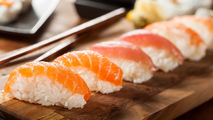 Mehrere Stücke Sushi mit Lachs und Thunfisch auf Holzbrett, daneben Stäbchen und unscharfe Hintergrunddetails.