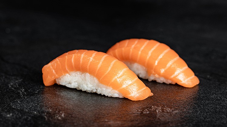 Zwei Stück Lachs-Nigiri mit Reis liegen auf einer schwarzen Steinplatte.