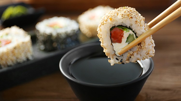 Ein Sushi-Rollstück mit Sesam wird mit Essstäbchen über eine schwarze Schale mit Sojasoße gehalten, weitere Sushi im Hintergrund.