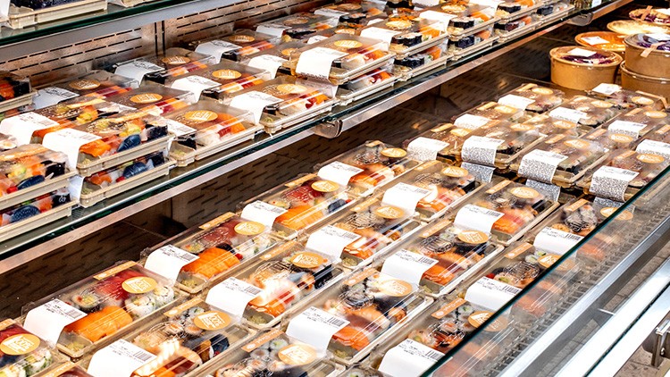 Kühlregal mit verpacktem Sushi in transparenten Plastikschalen und durchsichtigen Deckeln, teils mit Etiketten versehen.