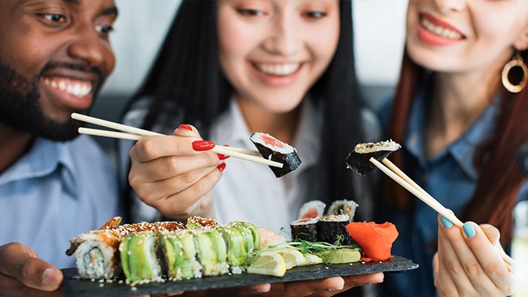 Mehrere Sushi-Rollen mit Avocado, Fisch und Sesam auf Schieferplatte, daneben Wasabi, Ingwer und Zitronenscheiben.